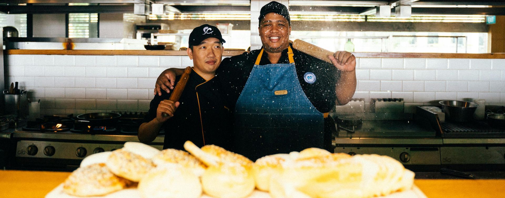 Two chefs in a restaurant kitchen with baked goods, smiling and holding a rolling pin.