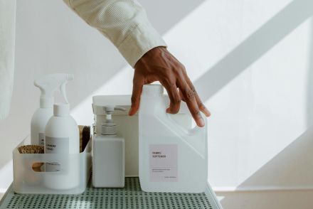 Close-up of a man holding fabric softener surrounded by cleaning products in a minimalist setting.